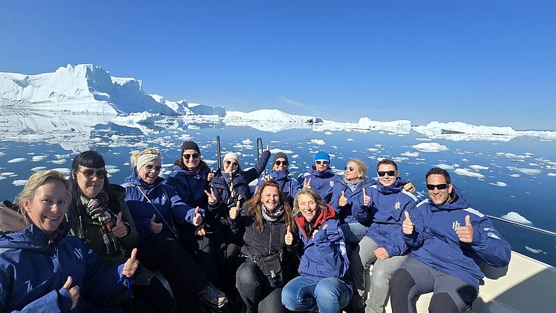 Eine Gruppe von Reiseprofis sitzt in blauen Expeditionsjacken auf einem Boot vor der beeindruckenden Kulisse von Eisbergen und schneebedeckten Landschaften in Westgrönland. Alle lächeln in die Kamera und zeigen Daumen hoch bei strahlend blauem Himmel.