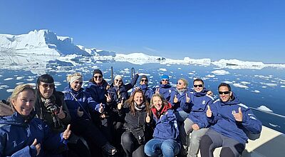 Eine Gruppe von Reiseprofis sitzt in blauen Expeditionsjacken auf einem Boot vor der beeindruckenden Kulisse von Eisbergen und schneebedeckten Landschaften in Westgrönland. Alle lächeln in die Kamera und zeigen Daumen hoch bei strahlend blauem Himmel.