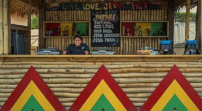 Schauinsland startet mit zehn Resorts auf Jamaika, hier einer der Foodhuts im Iberostar Waves Rose Hall Lächelnde dunkelhäutige Person in einer schön gestalteten Bambushütte, außerdem sind die jamaikanischen Nationalfarben rot, gelb, grün zu sehen. Die Hütte steht auf einem Hotelgelände