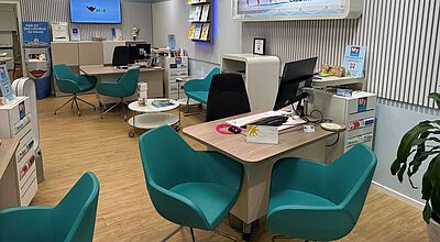 Ein modernes Reisebüro mit hellen Wänden, großen Werbetafeln für AIDA und Club Med, mehreren türkisfarbenen Sesseln und Beratungsplätzen mit Computern und Prospekten; die Atmosphäre ist freundlich und einladend.