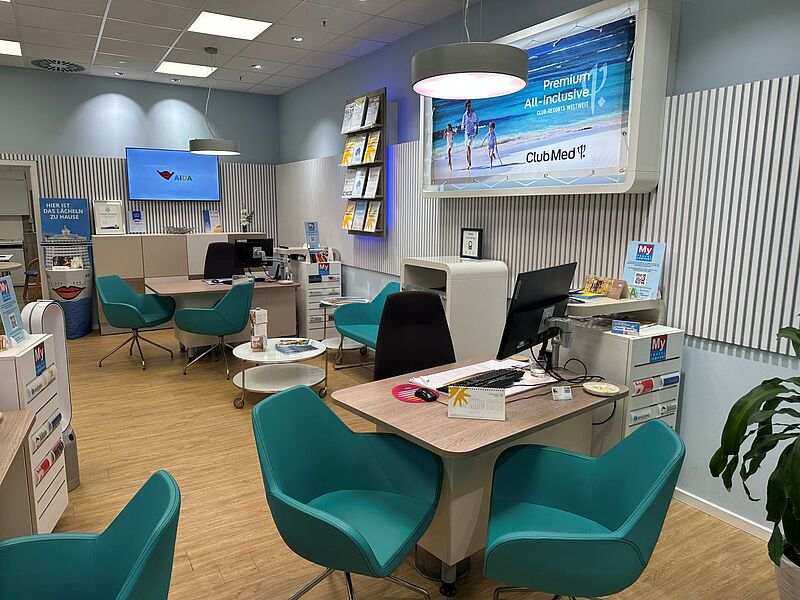 Ein modernes Reisebüro mit hellen Wänden, großen Werbetafeln für AIDA und Club Med, mehreren türkisfarbenen Sesseln und Beratungsplätzen mit Computern und Prospekten; die Atmosphäre ist freundlich und einladend.