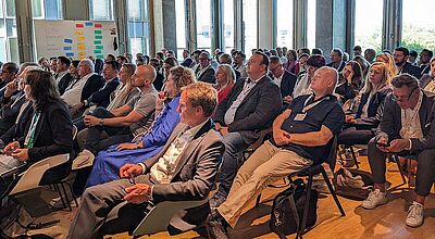 Ein Konferenzraum mit vielen sitzenden Teilnehmerinnen und Teilnehmern, die aufmerksam einem Vortrag folgen. Die Menschen sind überwiegend in Businesskleidung gekleidet. Im Hintergrund ist eine Pinnwand mit farbigen Zetteln sichtbar, die vermutlich Workshop-Themen zeigen. Der Raum ist hell, mit großen Fenstern und moderner Ausstattung.