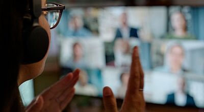 Eine Person mit Kopfhörern und Brille nimmt an einer Videokonferenz teil. Auf dem unscharfen Bildschirm im Hintergrund sind mehrere Teilnehmende in kleinen Fenstern zu sehen. Die Person im Vordergrund hebt gestikulierend die Hände, was auf eine aktive Beteiligung am Gespräch hindeutet.