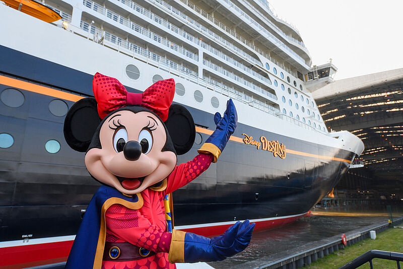 Eine mannshohe Minnie Maus in Superheldinnen-Kostüm steht vor dem neuen Kreuzfahrtschiff Disney Destiny und zeigt auf den dunklen Bug des Neubaus, der aus der Werfthalle gezogen und erstmals der Öffentlichkeit präsentiert wurde. 