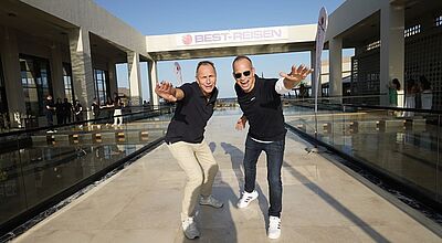 Dynamisches Duo: Die Best-Reisen-Vorstände Frank Winkler (links) und Cornelius Meyer am Eingang des Tagungshotels Aldiana Naga Bay in Ägypten. Foto Best-Reisen/Jörg Sänger Zwei Männer mittleren Alters im Freizeitlook zeigen sich dynamisch vor dem Eingang eines Tagungshotels.