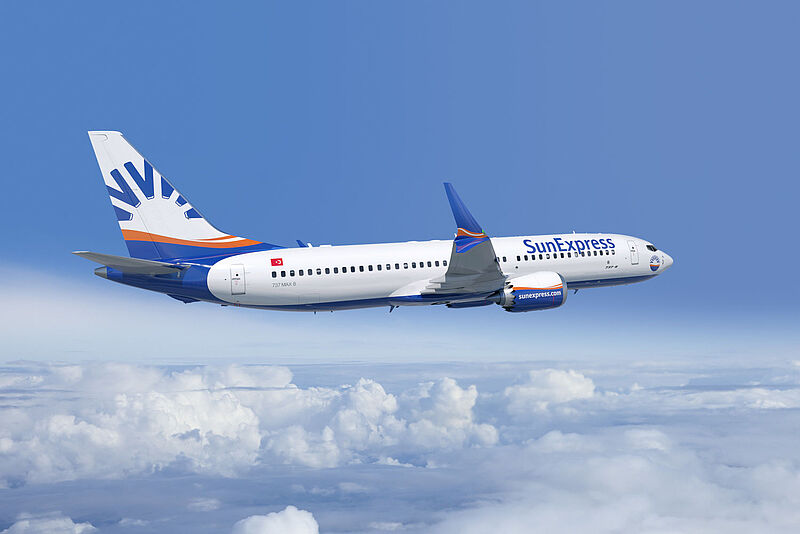 Das Bild zeigt ein Flugzeug der Fluggesellschaft SunExpress in der Luft. Es handelt sich um eine Boeing 737 MAX 8 mit dem charakteristischen blauen, weißen und orangefarbenen Design von SunExpress. Der Name der Airline ist auf dem Rumpf zu sehen, ebenso wie die Webadresse "sunexpress.com". Die türkische Flagge ist in der Nähe der hinteren Tür zu erkennen. Das Flugzeug fliegt über einer Wolkendecke vor einem klaren blauen Himmel.        4o