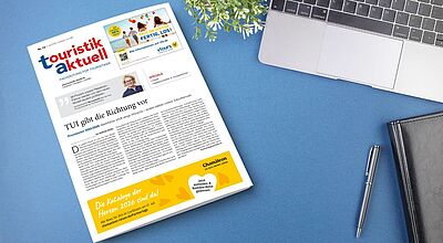 Auf einem blauen Schreibtisch liegt eine Ausgabe der Fachzeitung „touristik aktuell“ mit der Titelgeschichte „TUI gibt die Richtung vor“. Neben der Zeitung befinden sich ein silberner Laptop, ein schwarzes Notizbuch, ein silberner Kugelschreiber und eine grüne Pflanze. Am unteren Rand der Zeitung ist eine gelbe Anzeige von Chamäleon Reisen mit dem Text „Die Kataloge der Herzen 2026 sind da!“ zu sehen.