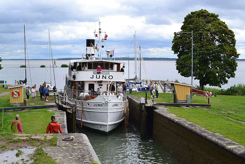 Von einem See im Süden Schwedens fährt das historische Passagierschiff Juno in eine enge Schleusentreppe des Göta-Kanals ein. Bestaunt wird das Manöver von Passagieren an Deck und an Land stehenden Schaulustigen.