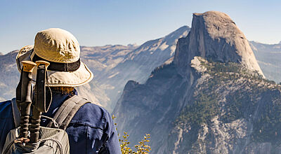 Eine Wandertouristin mit Rucksack und Wanderstöcken steht im Yosemite-Nationalpark und betrachtet den Half Dome, der im Hintergrund unscharf zu sehen ist.