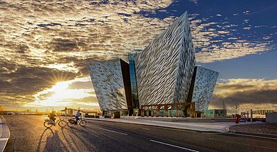 Modernes Gebäude des Titanic Museums in Belfast bei Sonnenuntergang, mit zwei Radfahrern auf einer breiten Straße im Vordergrund
