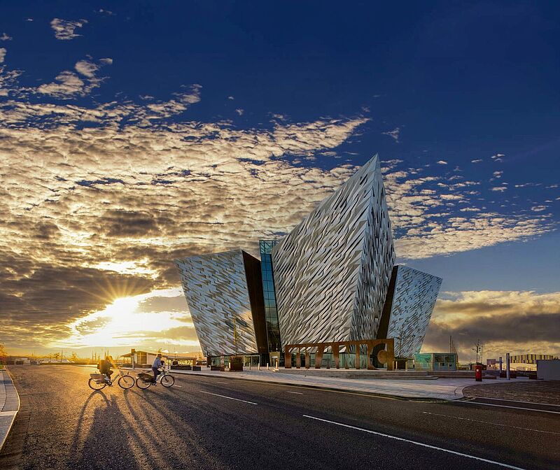 Modernes Gebäude des Titanic Museums in Belfast bei Sonnenuntergang, mit zwei Radfahrern auf einer breiten Straße im Vordergrund