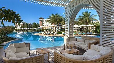 Luxuriöse Poolanlage mit weißer Pergola-Lounge, Liegestühlen, Sonnenschirmen und Palmen vor mediterraner Hotelarchitektur mit Turm.