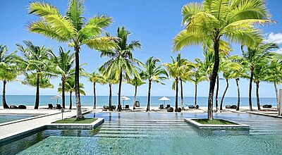 Tropisches Strandresort: Infinity-Pool umgeben von Palmen, im Hintergrund türkisblaues Meer und weißer Sandstrand.