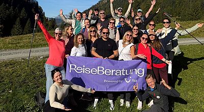 Die TLTU-Berater waren in diesem Jahr viel unterwegs Eine große Gruppe fröhlicher Menschen posiert im Grünen vor einer Bergkulisse. Sie halten ein lilafarbenes Banner mit der Aufschrift „Reiseberatung – Powered by TUI“. Viele heben lachend die Arme, einige knien oder stehen davor. Der Himmel ist klar und blau, die Umgebung wirkt wie eine alpine Landschaft.