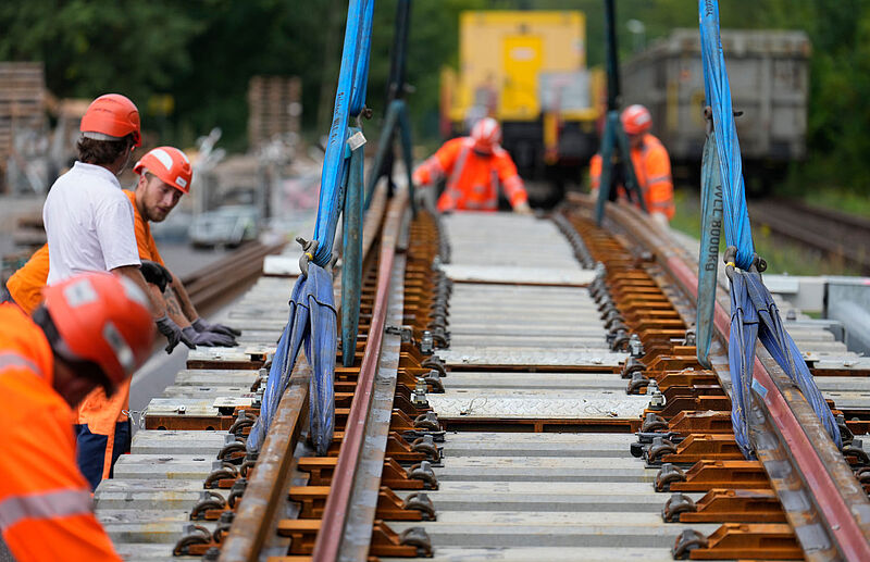 Bauarbeiter in orangefarbener Schutzkleidung verlegen neue Bahngleise mit Hilfe von blauen Hebevorrichtungen.