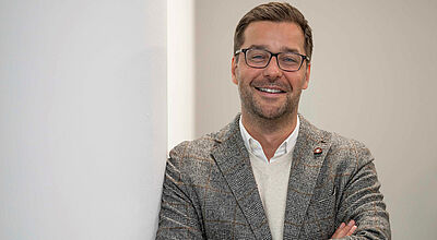 Lächelnder Mann mit Brille in grauem Jackett und weißem Hemd, verschränkte Arme, an weißer Wand lehnend.