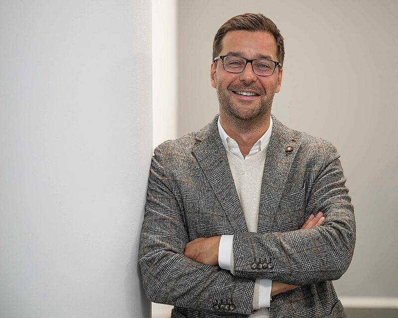 Lächelnder Mann mit Brille in grauem Jackett und weißem Hemd, verschränkte Arme, an weißer Wand lehnend.