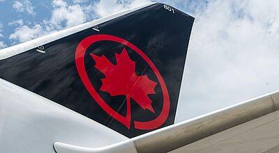 Nahaufnahme eines Seitenleitwerks auf einem Flugzeug von Air Canada mit schwarz-rotem Ahorn-Logo vor wolkigem Himmel.