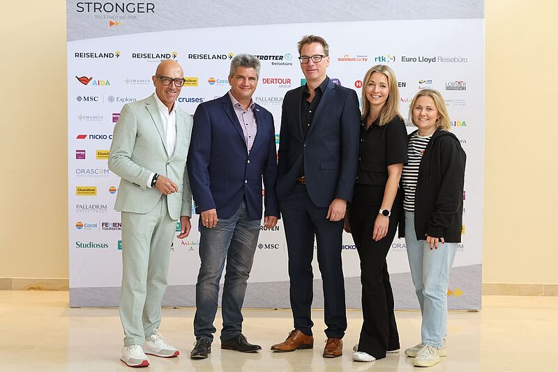 Drei Männer und zwei Frauen posieren in einem Innenraum vor einer Fotowand auf der viele Logos abgebildet sind und lächeln zum Fotografen in die Kamera.