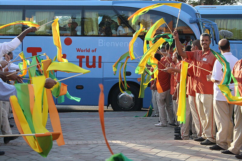 TUI ist bereit für den Sommer 2026: Vor allem Familien sollen mit Sonderangeboten als Frühbucher gewonnen werden. Mehrere Menschen in Reiseleiter-Kleidung stehen einander gegenüber und schwenken gelbe, grüne und orangefarbene Stoffbänder, während im Hintergrund ein hellblauer Reisebus mit der Aufschrift „World of TUI“ ankommt.