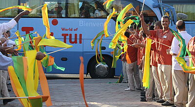 TUI ist bereit für den Sommer 2026: Vor allem Familien sollen mit Sonderangeboten als Frühbucher gewonnen werden. Mehrere Menschen in Reiseleiter-Kleidung stehen einander gegenüber und schwenken gelbe, grüne und orangefarbene Stoffbänder, während im Hintergrund ein hellblauer Reisebus mit der Aufschrift „World of TUI“ ankommt.