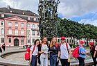 Vier gut gelaunte Personen stehen vor einem kunstvoll gestalteten Brunnen mit vielen kleinen Bronzefiguren, dem Fastnachtsbrunnen in Mainz. Sie tragen weiße oder dunkle Oberteile und rote Rucksäcke, zwei von ihnen auch rote „Emirates“-Kappen. Im Hintergrund ist ein barockes Gebäude mit rosafarbener Fassade und weiteren Teilnehmerinnen mit roten Kappen zu sehen. Die Gruppe wirkt entspannt und fröhlich, vermutlich bei einer Stadtrallye oder Gruppenveranstaltung. Der Platz ist gepflastert und von Bäumen und historischen Gebäuden umgeben.