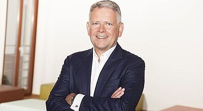 Ein lächelnder Mann mittleren Alters mit grauem Haar trägt ein dunkelblaues Sakko und ein weißes Hemd. Er steht mit verschränkten Armen in einem modernen, hellen Büro mit farbigen Sitzmöbeln im Hintergrund.