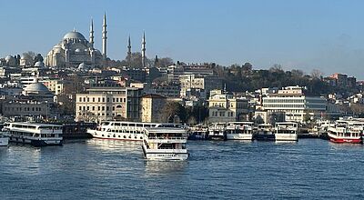 Blick über das Wasser des Goldenen Horns in Istanbul, Türkei, mit zahlreichen weißen und bunten Ausflugsbooten im Vordergrund. Im Hintergrund erhebt sich die imposante Süleymaniye-Moschee mit ihren hohen Minaretten über die dicht bebaute Altstadt. Der Himmel ist klar und blau, die Szene wirkt ruhig und sonnig.