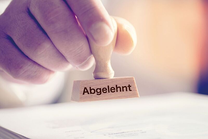 Hand drückt Holzstempel mit Aufschrift "Abgelehnt" auf Dokument, blaue und rosa Beleuchtung.