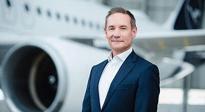 Lufthansa-Airlines-Chef Jens Ritter ist mit dem „Turnaround“-Programm bislang „äußerst zufrieden“ Ein Manager mittleren Alters im dunkelblauen Anzug und weißen Hemd steht vor einem Flugzeug von Lufthansa, das sich unscharf im Hintergrund befindet.