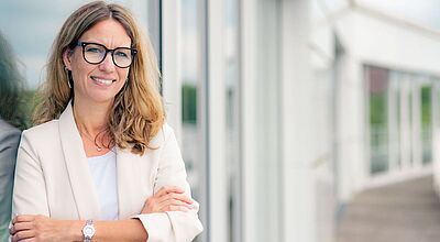 Eine Frau mit langen, blonden Haaren steht lächelnd mit verschränkten Armen vor einem modernen Gebäude mit Glasfassade. Sie trägt eine helle Blazerjacke, eine Brille mit dunklem Rahmen und eine Armbanduhr. Die Atmosphäre wirkt professionell und freundlich, passend zu einem Business- oder Porträtfoto.