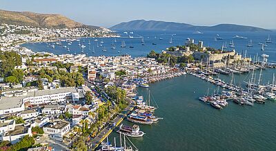 Luftaufnahme des türkischen Küstenorts Bodrum mit seinem Yachthafen, zahlreichen Booten in der türkisfarbenen Bucht, weißen Häusern entlang der Uferpromenade und der markanten Burg von Bodrum im Hintergrund.