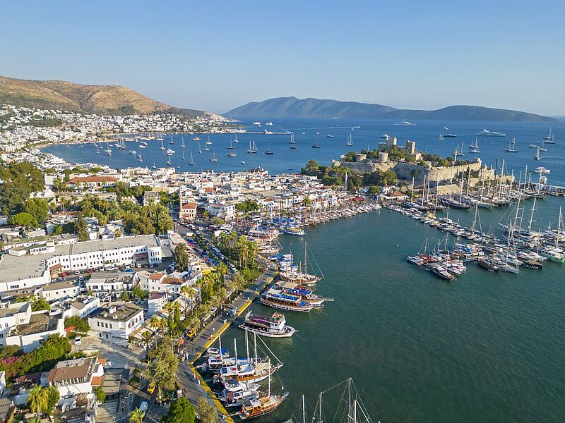 Luftaufnahme des türkischen Küstenorts Bodrum mit seinem Yachthafen, zahlreichen Booten in der türkisfarbenen Bucht, weißen Häusern entlang der Uferpromenade und der markanten Burg von Bodrum im Hintergrund.