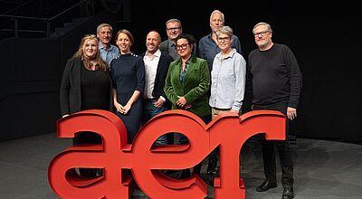 Eine Gruppe von neun Personen steht gemeinsam auf einer Bühne hinter einem großen, roten 3D-Logo mit den Buchstaben „aer“. Die Personen lächeln in die Kamera. Der Hintergrund ist dunkel, und im oberen Bereich ist teilweise eine Treppe zu sehen.