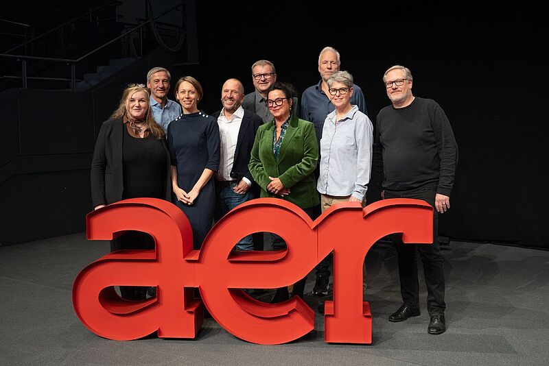 Eine Gruppe von neun Personen steht gemeinsam auf einer Bühne hinter einem großen, roten 3D-Logo mit den Buchstaben „aer“. Die Personen lächeln in die Kamera. Der Hintergrund ist dunkel, und im oberen Bereich ist teilweise eine Treppe zu sehen.