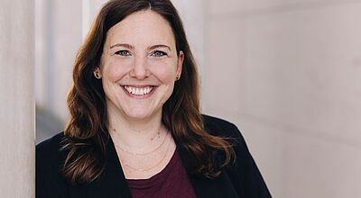 Porträt einer lächelnden Frau mit schulterlangen braunen Haaren. Sie trägt eine dunkle Jacke über einem bordeauxfarbenen Shirt und steht vor einem hellen, unscharfen Hintergrund.