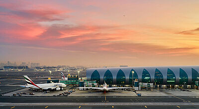 Flughafen in Dubai: bislang keine Streichungen von Emirates und deutschen Airlines Das moderne Terminal des Flughafens Dubai bei Sonnenuntergang mit türkis verglasten Bögen, Emirates-Flugzeugen am Gate und einem dramatischen rosa-goldenen Himmel.