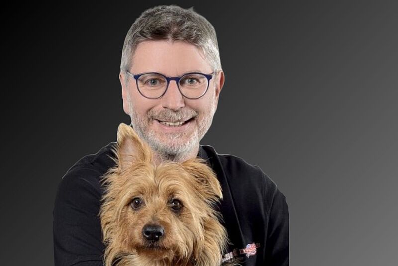 Mann mit schwarzem Pulli, Brille und grauen Haaren und Bart lächelt in die Kamera, ein kleiner, brauner Hund auf seinem Schoß. 