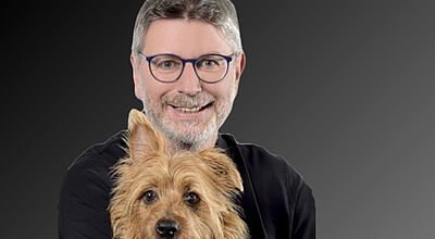Werner Salheiser mit seinem Hund Murphy. Mann mit schwarzem Pulli, Brille und grauen Haaren und Bart lächelt in die Kamera, ein kleiner, brauner Hund auf seinem Schoß.