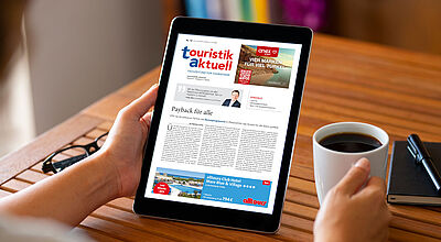 Eine Person hält ein Tablet in der linken Hand, auf dem die digitale Ausgabe der Fachzeitung touristik aktuell angezeigt wird. In der rechten Hand hält die Person eine weiße Kaffeetasse. Auf dem Tablet ist ein Artikel mit dem Titel „Payback für alle“ zu sehen sowie Werbeanzeigen für Reiseangebote. Im Hintergrund liegt eine Brille auf dem Holztisch, daneben ein Notizbuch mit einem Stift.