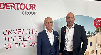 Zwei Männer in Anzügen stehen vor einer Wand mit Dertour-Werbung