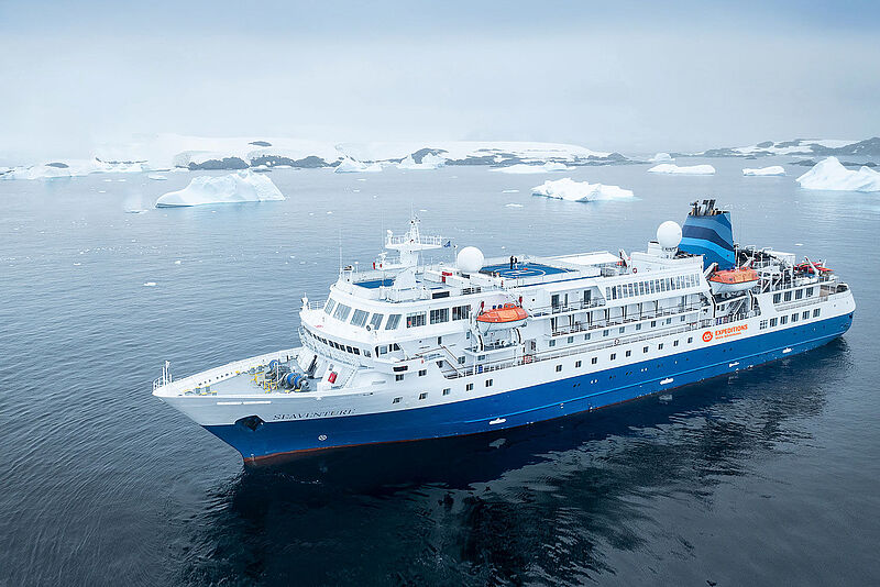 Ein Expeditionskreuzfahrtschiff mit blauem Rumpf liegt in einer Polarregion, im Hintergrund treiben flache Eisberge