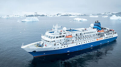 Die Seaventure, ex Bremen, ist nun wieder auf dem deutschen Markt buchbar. Foto: 66° Expedition Cruise Ein Expeditionskreuzfahrtschiff mit blauem Rumpf liegt in einer Polarregion, im Hintergrund treiben flache Eisberge