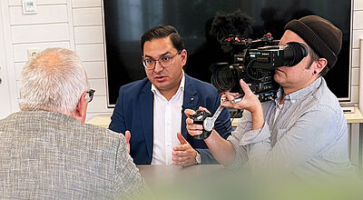 Schmetterling-Geschäftsführer Ömer Karaca forderte im Interview, die Wertigkeit der Frühbucherrabatte zu stärken Ein Mann mit schwarzem Jacket und weißem Hemd sitzt mit zwei weiteren Männern an einem Tisch. Ein Mann mit grauen Haaren sitzt mit dem Rücken zum Fotografen, ein anderer hält eine Kamera in der Hand und filmt die Situation.