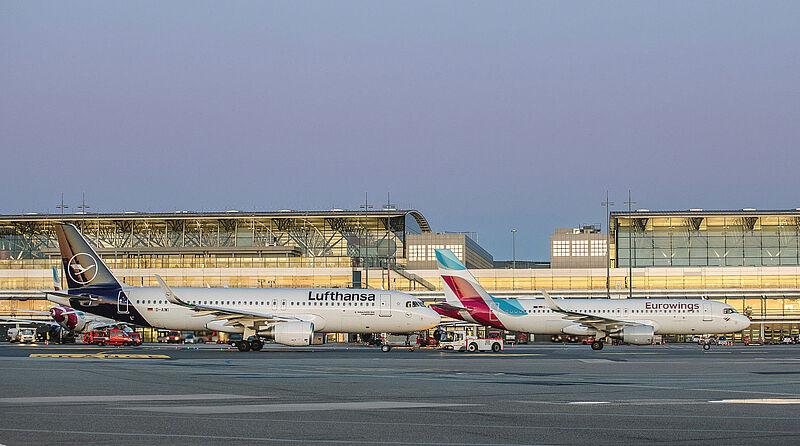 Lufthansa und Eurowings planen erst einmal keine zusätzliche Ausdünnung des Inlandsnetzes Flugzeuge von Lufthansa und Eurowings stehen vor dem Terminalgebäude des Hamburger Flughafens, angeleuchtet von der Abendsonne.