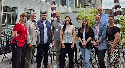 „Eine Gruppe von neun Personen steht lächelnd auf einer Dachterrasse in einer Stadt, im Hintergrund sind moderne Hochhausgebäude mit Glasfassade zu sehen. Die Gruppe trägt Namensschilder und wirkt wie bei einem geschäftlichen oder branchenspezifischen Treffen.