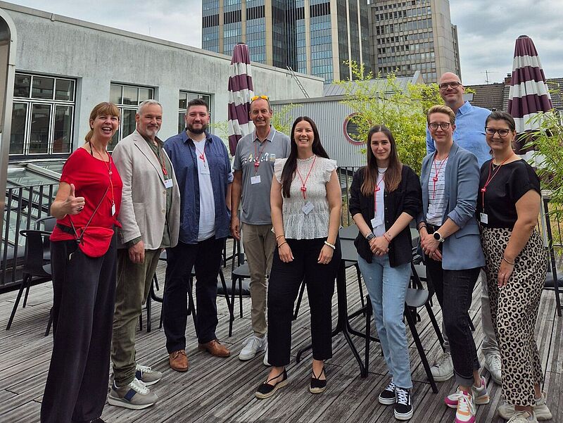 „Eine Gruppe von neun Personen steht lächelnd auf einer Dachterrasse in einer Stadt, im Hintergrund sind moderne Hochhausgebäude mit Glasfassade zu sehen. Die Gruppe trägt Namensschilder und wirkt wie bei einem geschäftlichen oder branchenspezifischen Treffen.