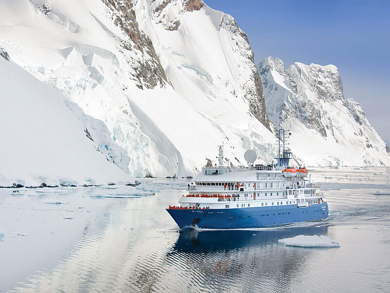 Ein kleines Expeditionskreuzfahrtschiff mit blauem Rumpf und an Deck stehenden Passagieren in roten Anoraks passiert einen hohen Eisberg