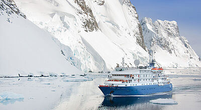 Das kleine Expeditionsschiff Sea Spirit reist mit maximal 114 Passagieren ins Eis. Foto: Poseidon Expeditions Ein kleines Expeditionskreuzfahrtschiff mit blauem Rumpf und an Deck stehenden Passagieren in roten Anoraks passiert einen hohen Eisberg