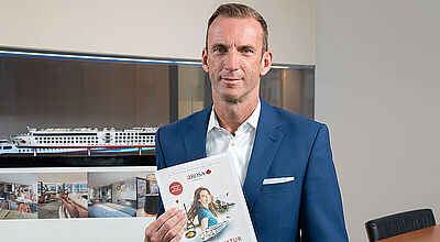 Arosa-Vertriebschef Stefan Sprunk berichtet von guten Vorausbuchungen für 2025. Foto: Arosa Arosa-Vertriebschef Stefan Sprunk mit Katalog 2025 vor Schiffsmodelloto: Arosa